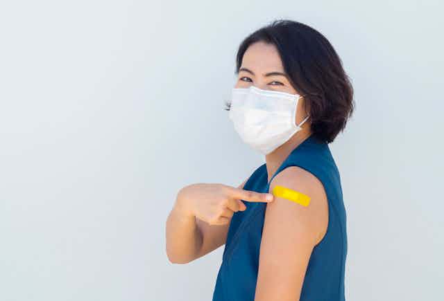 Woman in a face mask pointing to a bandage on her arm after getting vaccinated