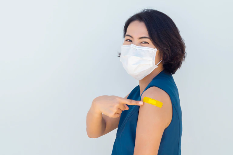 Woman in a face mask pointing to a bandage on her arm after getting vaccinated