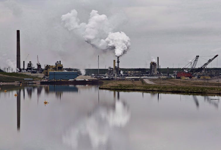 The Syncrude oil sands extraction facility is seen, reflected in a body of water in front of it.