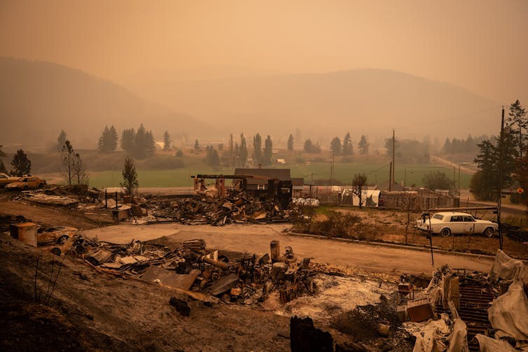 Thick orange-ish smoke fills the air and blocks out the sun over a neighbhourhood destroyed by a wildfire.