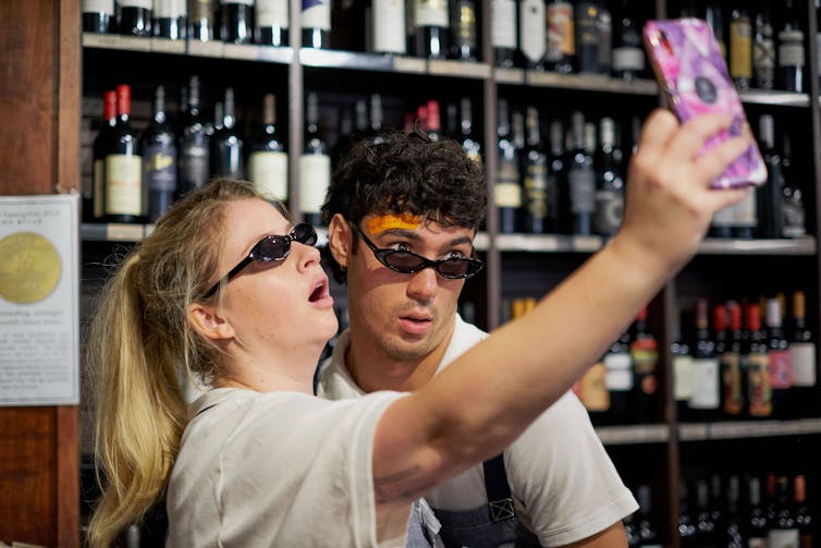 A man and a woman pose for a selfie in a bottle shop, both wearing pointy sunglasses