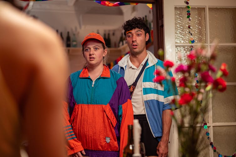 A man and a woman in brightly-coloured windcheaters, staring in silent awe into a party scene