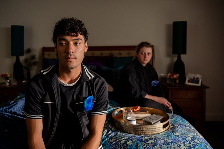 A man sits on the edge of a bed looking ahead with a serious expression. Behind him, a woman sits in the shadows