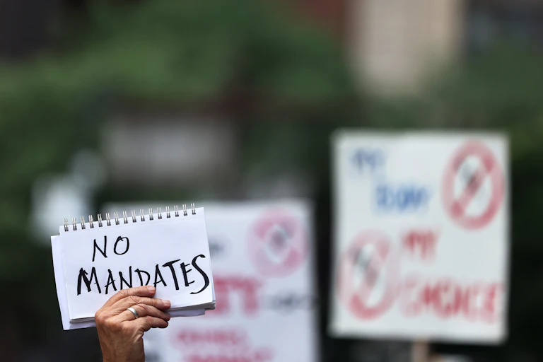 A hand with a silver ring on its fourth finger holds up a white spiral-bound piece of paper with the words "No mandates" written on it in black. Other placards are visible in the background, though they are blurred and unreadable.