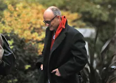 A bald man in a black coat and red scarf walks with his head down.