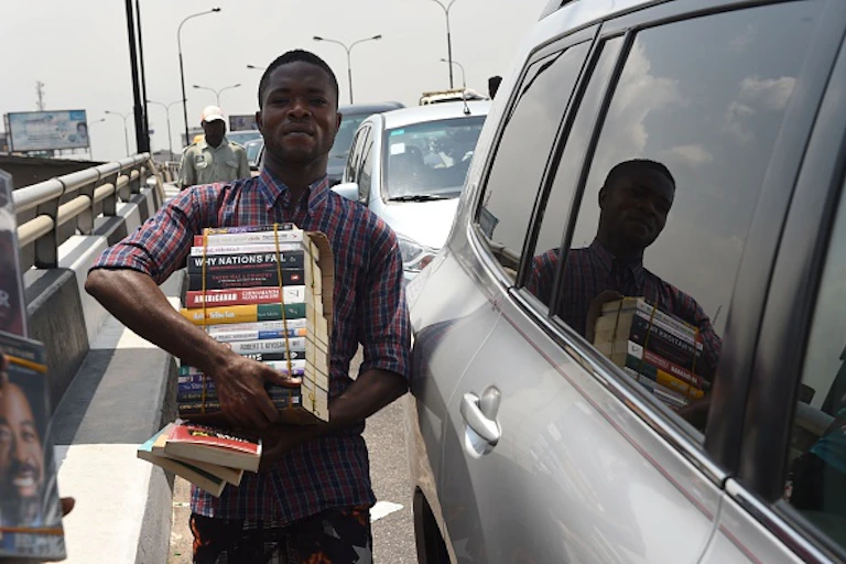 Young man carrying a pile of books stands next to a car on a road