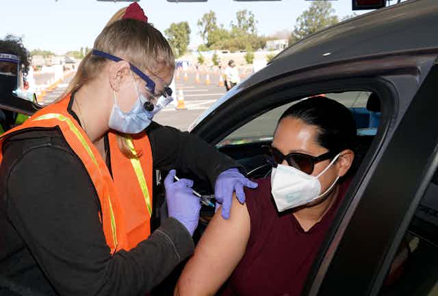 A woman wearing sunglasses and a white mask sitting in a car receives a vaccine shot from another woman standing and wearing protective gear