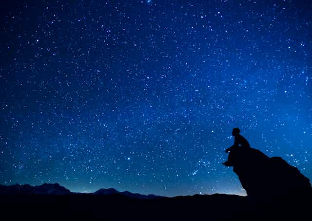 A person sits on a rock promontory against a vast night sky full of stars.