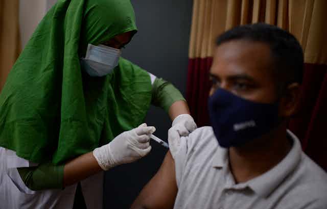 A man wearing a mask receives a shot of vaccine from a woman wearing a green head scarf in Dhaka, Bangladesh