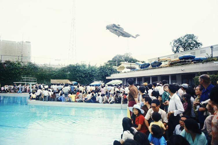 A crowd of people surrounds a swimming pool while a helicopter flies overhead