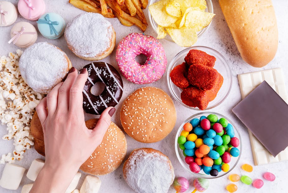 A woman's hand reaches for a donut, which is nestled among other sugary foods on a table, including candies and bread.