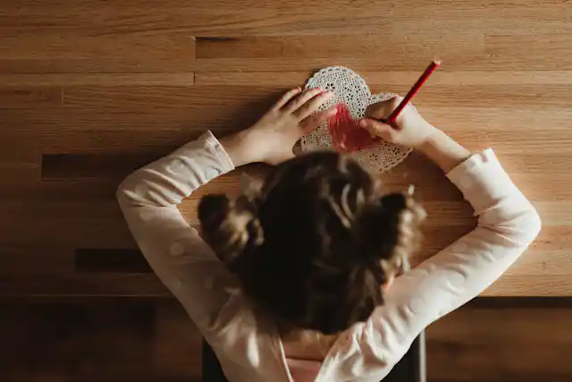 A girl draws on a heart-shaped card with a red coloring pencil