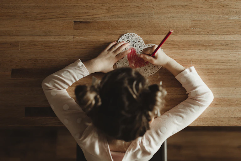 A girl draws on a heart-shaped card with a red coloring pencil
