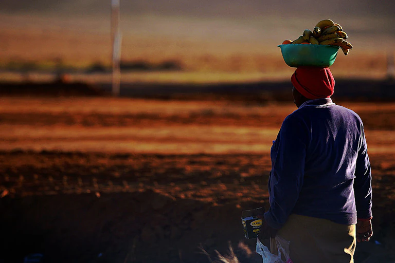 Sunset and a woman walks away from camera, a plastic bowl of fruit balanced on her head.