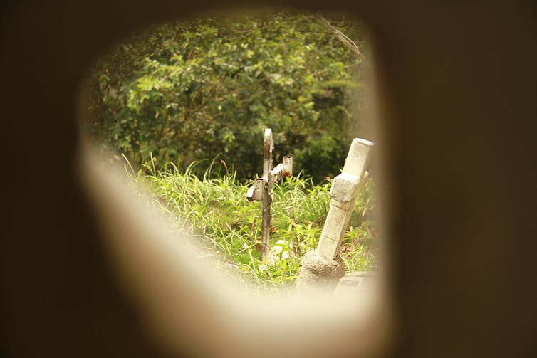 A view through an opening shows an overgrown field with two crosses leaning at angles.