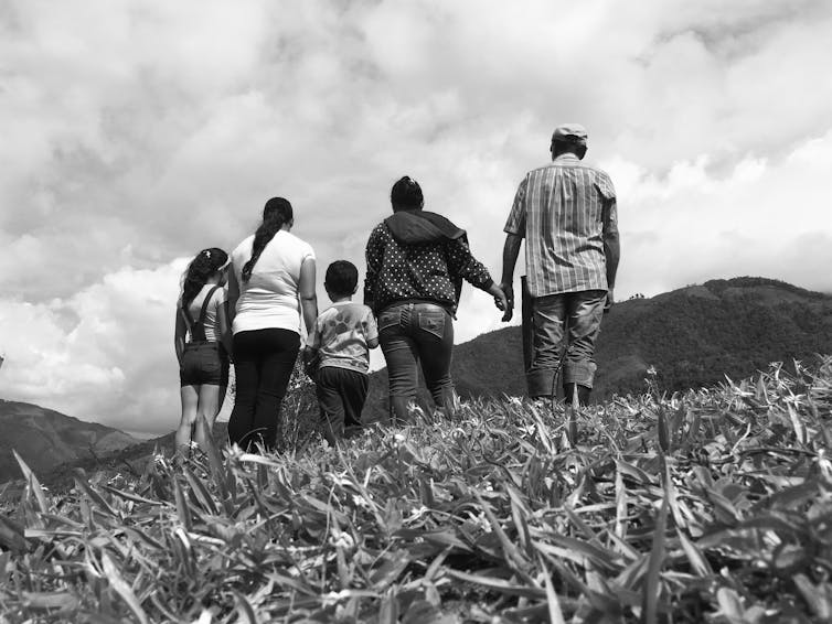 A black-and-white image of people of multiple ages standing in a field, holding hands, with their backs to the camera
