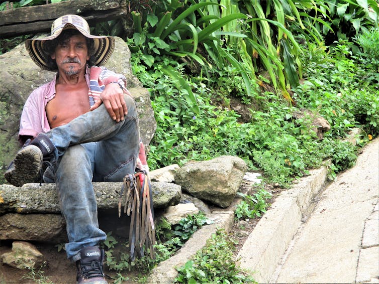 A man in a floppy hat, open shirt, jeans and boots sits on a rock next to greenery