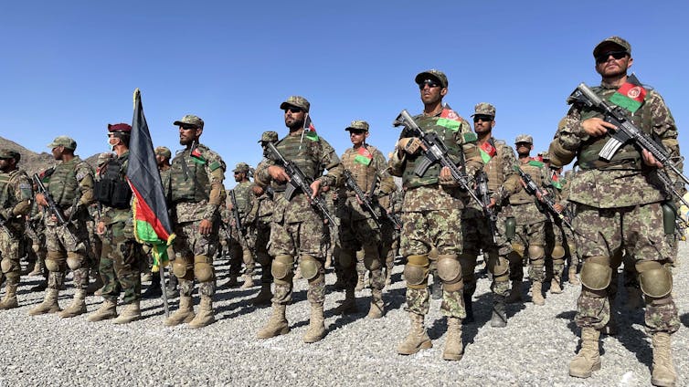 A group of people in camouflage uniforms carry rifles while standing in a formation behind the Afghan flag.