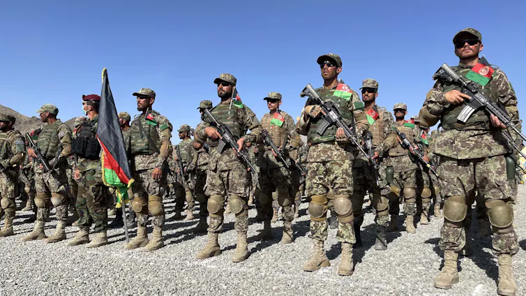 A group of people in camouflage uniforms carry rifles while standing in a formation behind the Afghan flag.