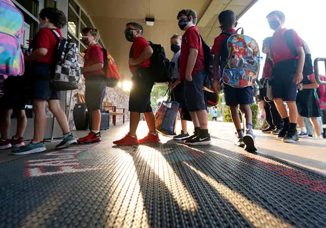 Young children lining up to enter school, all wearing masks and carrying backpacks.