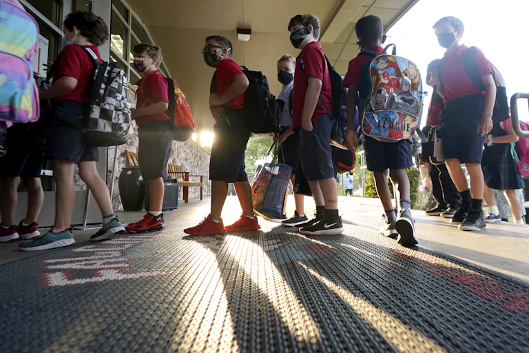Young children lining up to enter school, all wearing masks and carrying backpacks.