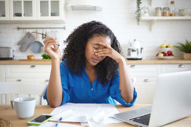 African-Australian woman working from her kitchen table puts her hand to her face, looking exhausted.