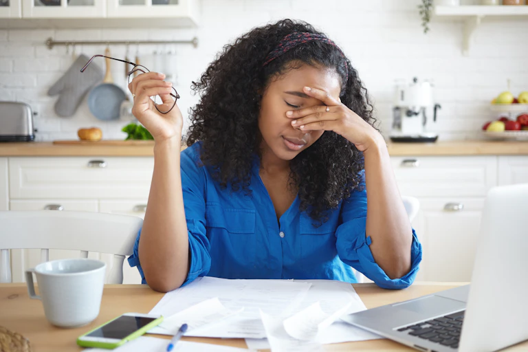African-Australian woman working from her kitchen table puts her hand to her face, looking exhausted.