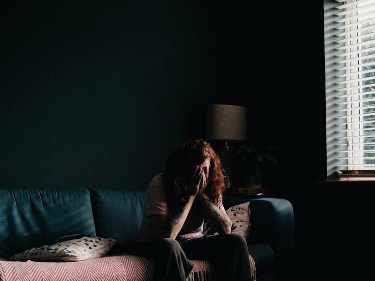 Person sits in a dark room on the couch with their head in their hands.