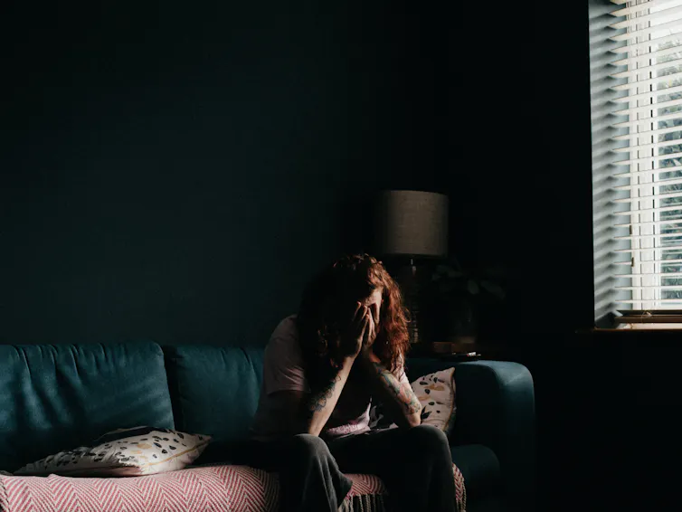 Person sits in a dark room on the couch with their head in their hands.