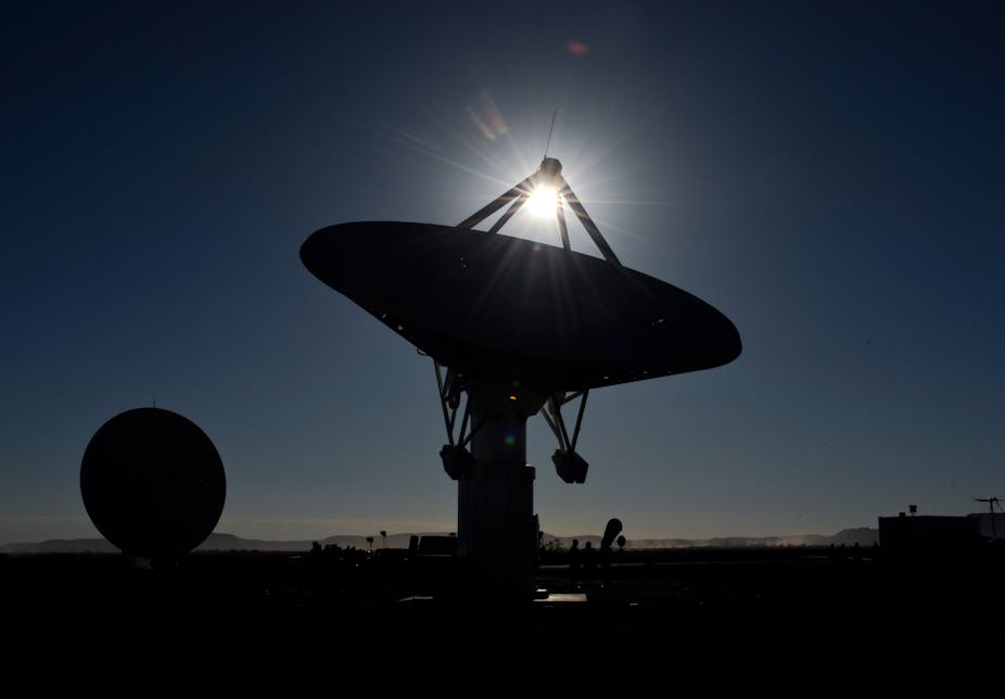 A huge satellite dish is pointed up at a darkening sky as the sun descends past it. Another similar dish is visible in the background.