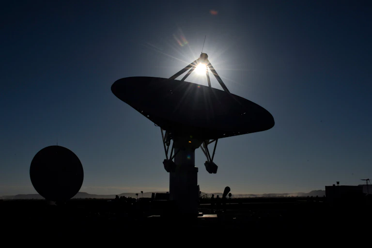 A huge satellite dish is pointed up at a darkening sky as the sun descends past it. Another similar dish is visible in the background.