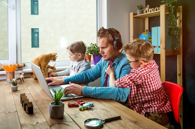 A busy dad homeschools his two children, while working on his laptop.