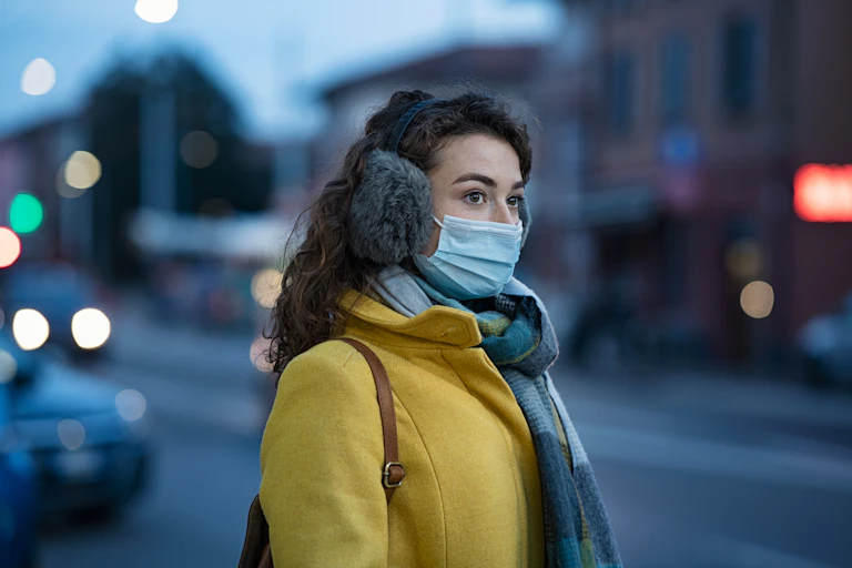 A woman in a mask and ear warmers looks down the road at dusk.