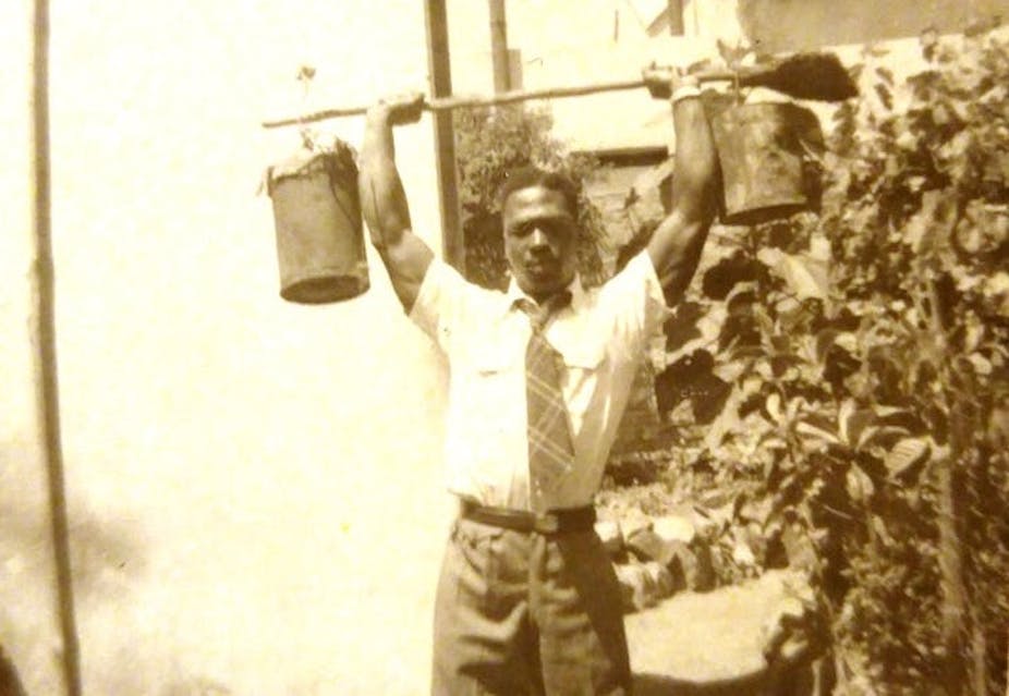 Man wearing long trousers, shirt and tie, lifting a gardening implement above his head with buckets suspended at each end.