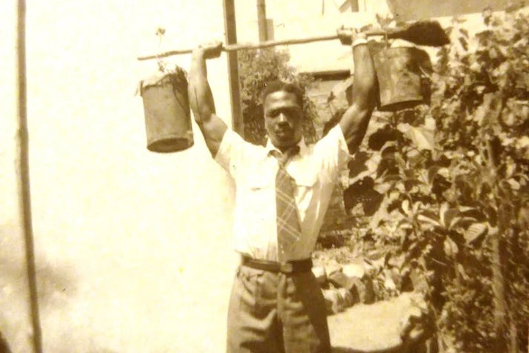 Man wearing long trousers, shirt and tie, lifting a gardening implement above his head with buckets suspended at each end.