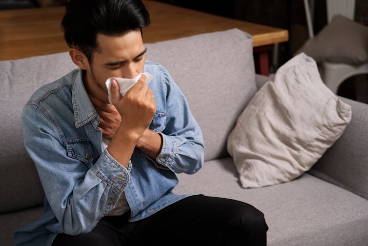 A man sits on a couch, coughing into a tissue.