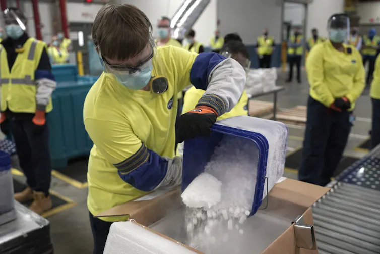 Un homme vêtu d’un équipement de protection et d’un tee-shirt jaune verse de la neige carbonique dans un carton contenant les vaccins Pfizer