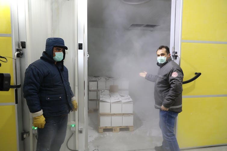 Two men stand next to a cold storage warehouse that contains boxes of Pfizer vaccine.