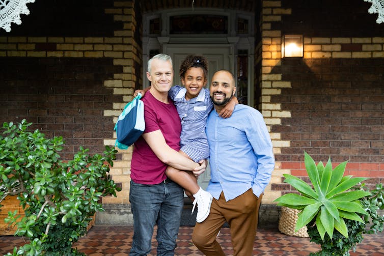 Two fathers stand in front of home with daughter holding backpack