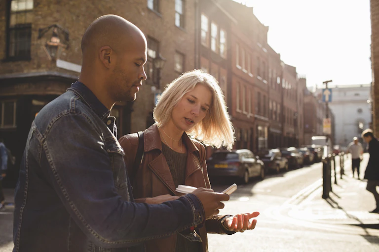 A man and a woman stand on a city street. The man holds a phone so that the woman can see it. She holds her left hand palm up.