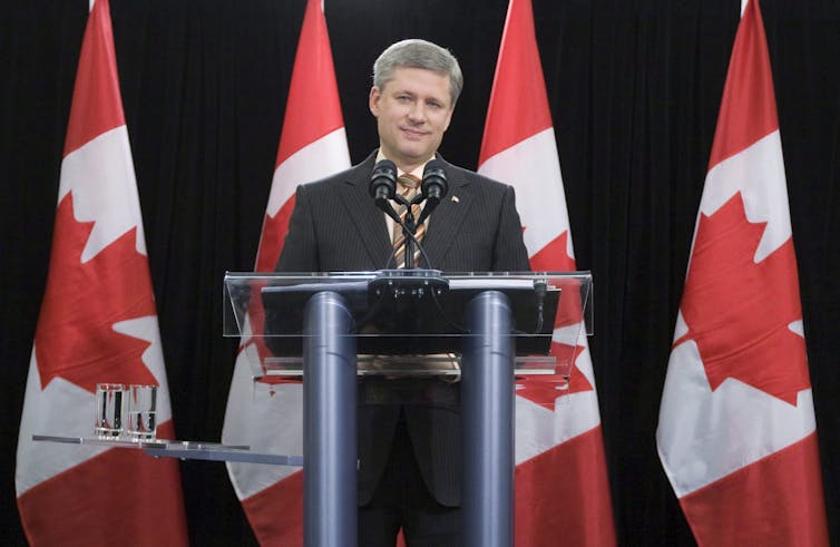 Stephen Harper stands at a podium with Canadian flags behind him.