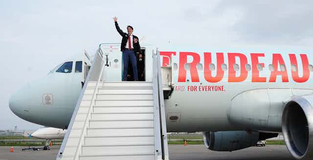 Trudeau is waving from the top of the stairs as he prepares to board his campaign plane that has the word TRUDEAU painted on it.