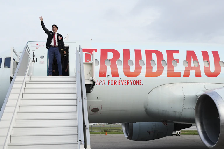 Trudeau is waving from the top of the stairs as he prepares to board his campaign plane that has the word TRUDEAU painted on it.