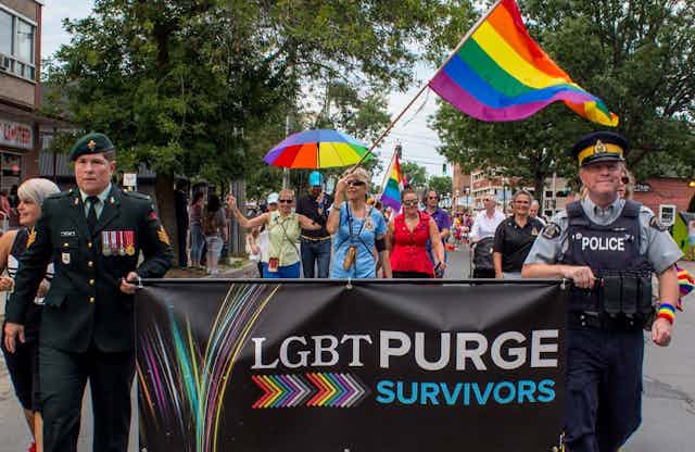 A military officer and police carry a sign that reads 'LGBT PURGE SURVIVORS' during a Pride Parade