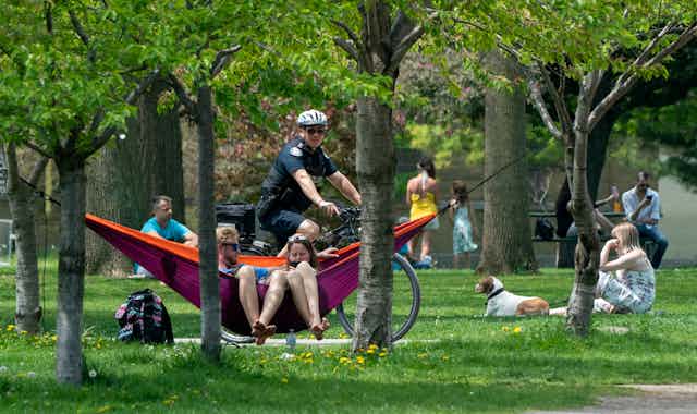 A bicycle police officer patrols Trinity Bellwoods Park. He is seen biking behind a couple in a hammock and a woman with her dog sit in the background.