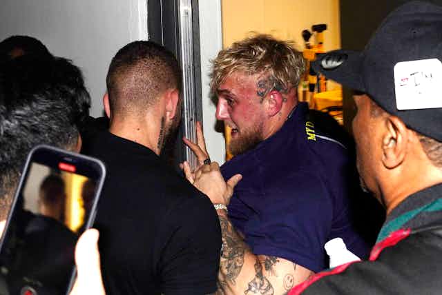 Jake Paul's face is very red as he is being pushed into a closet. People surround the influencer
