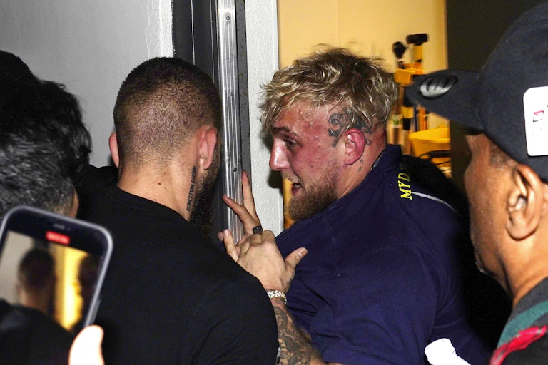 Jake Paul's face is very red as he is being pushed into a closet. People surround the influencer