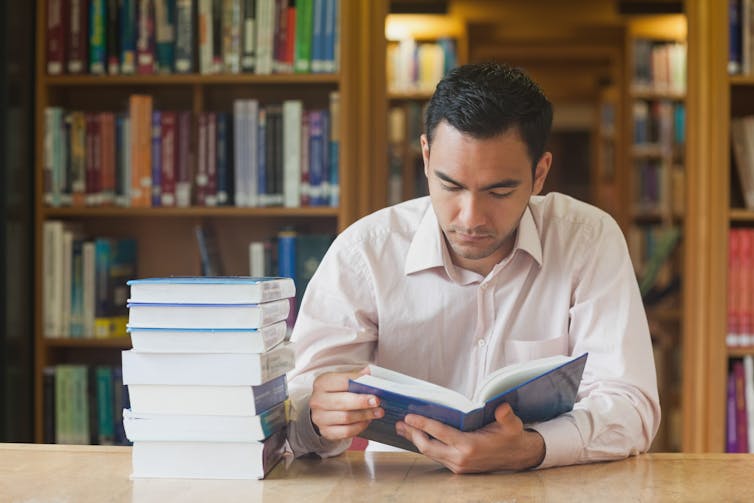 Male student reads a book with a pile of other books next to hhim