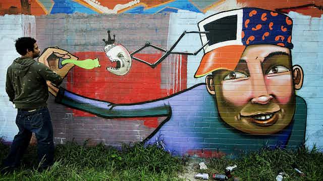 A graffittì artist works on a mural of a young 'coloured' man with a smile and a cap on his head with a door from which a face emerges on a spring to eat a green bug being held by the young man who is feeding it.