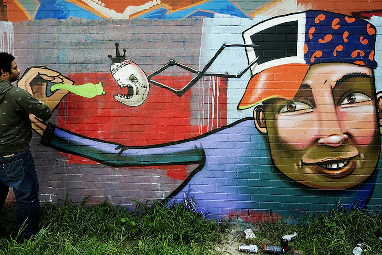 A graffittì artist works on a mural of a young 'coloured' man with a smile and a cap on his head with a door from which a face emerges on a spring to eat a green bug being held by the young man who is feeding it.